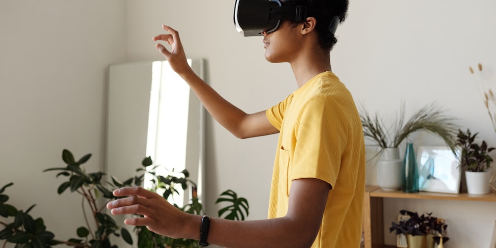 Młoda osoba w żółtej koszulce używająca gogli VR w jasnym pomieszczeniu z roślinami w tle.
