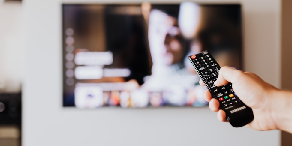 Ręka trzymająca pilota do telewizora z włączonym ekranem w tle
